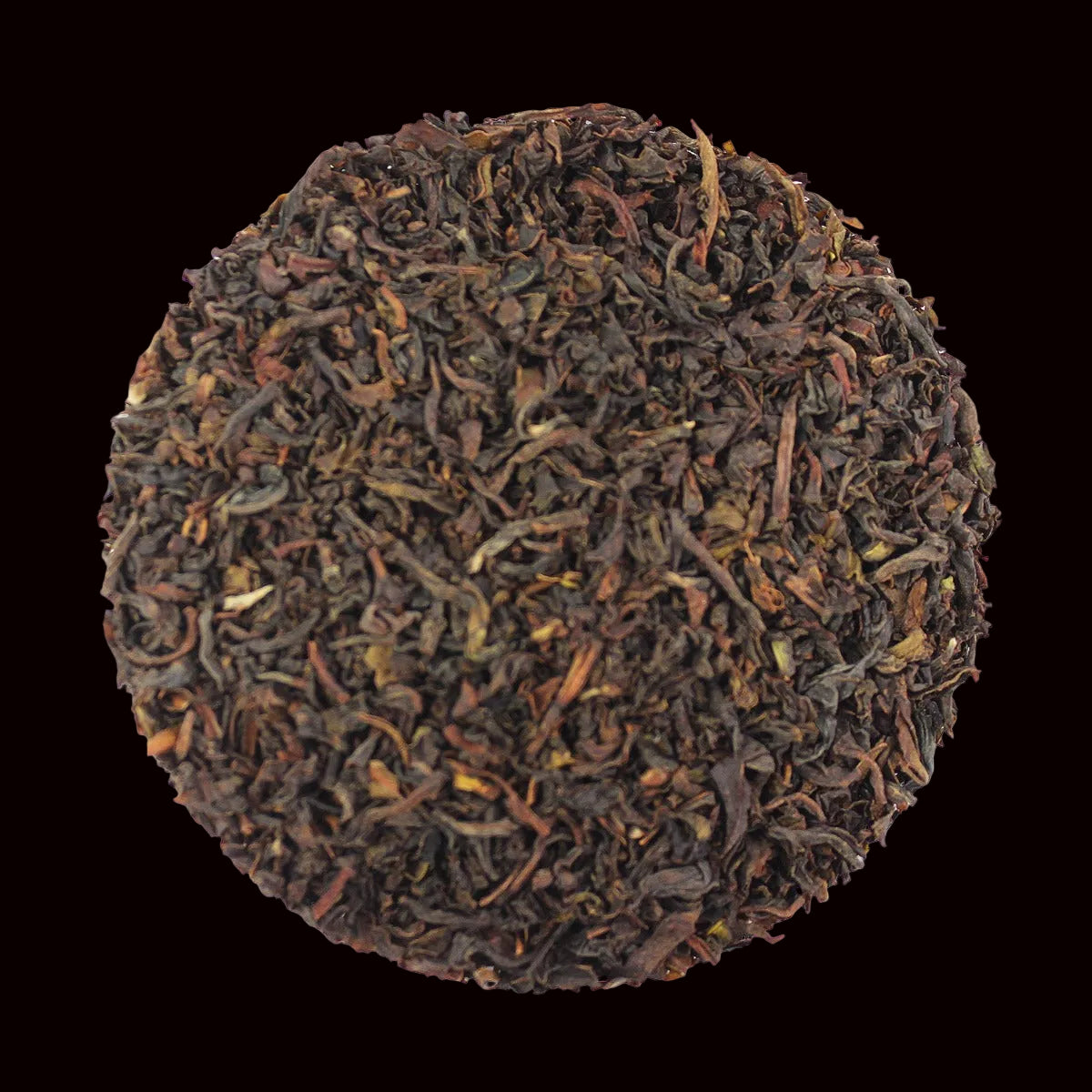 A circular pile of loose-leaf tea leaves, showing dark brown and reddish-brown tones with some lighter brown and yellowish-green tips, isolated on a black background.